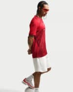 Athlete in a red sports jersey and white shorts, wearing red sunglasses and white‑red sneakers, standing in profile with hands behind back.