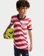 Boy wearing a USA soccer jersey with red and white stripes holding a bright yellow soccer ball by his side.