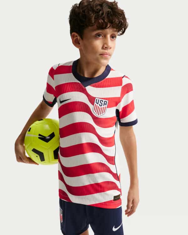 Boy wearing a USA soccer jersey with red and white stripes holding a bright yellow soccer ball by his side.
