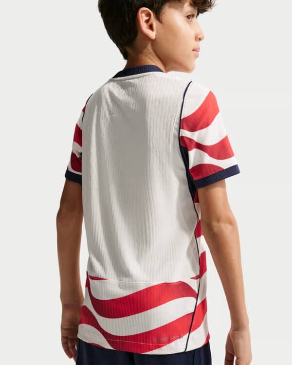 Back view of a boy wearing a white sports jersey with red wave patterns on the sleeves and hem, navy collar and cuffs.