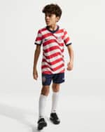Young boy wearing a USA soccer uniform with red and white striped shirt, navy shorts, white socks, and black cleats.