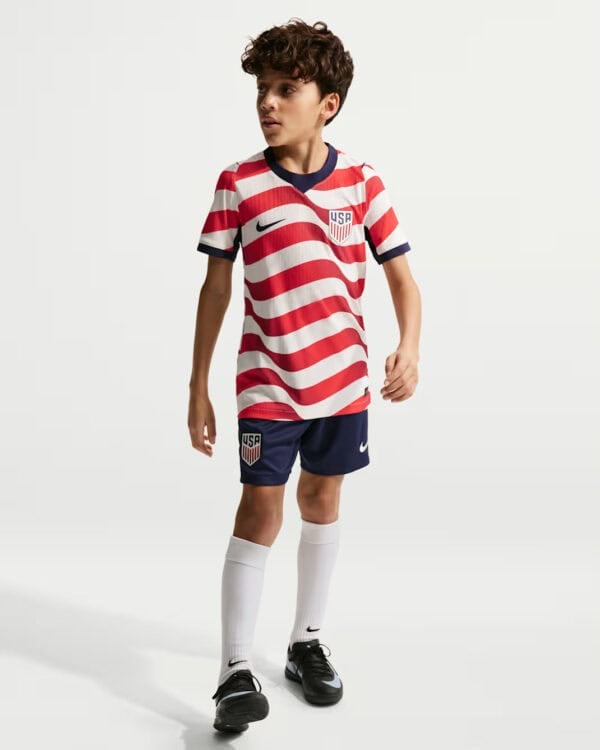 Young boy wearing a USA soccer uniform with red and white striped shirt, navy shorts, white socks, and black cleats.