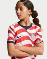Young woman wearing a USA soccer jersey with red and white wavy stripes, navy trim, and visible Nike swoosh and USA crest, arms crossed, looking to her left.