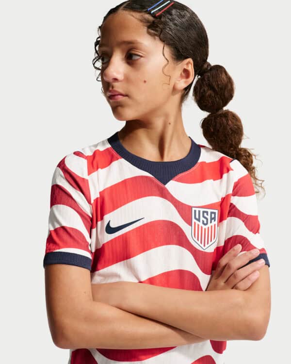 Young woman wearing a USA soccer jersey with red and white wavy stripes, navy trim, and visible Nike swoosh and USA crest, arms crossed, looking to her left.