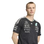 Portrait of a young man in a dark, geometric-patterned team jersey with sponsor logos, standing against a white background.