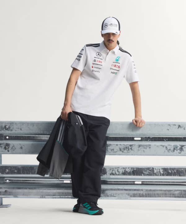 Man in a white racing polo and cap leaning on a metal guardrail, holding a black jacket.