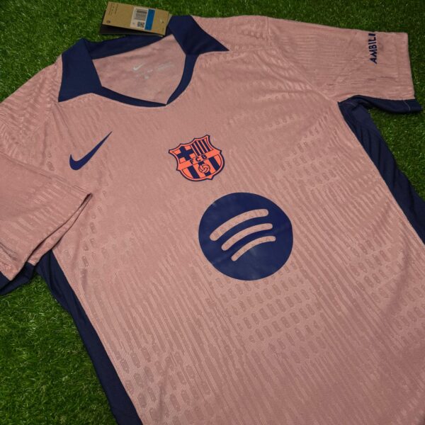 Pink FC Barcelona football jersey with navy collar and cuffs on grass, showing Nike swoosh and club crest with Spotify sponsor logo nearby.