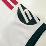 00143f52-923b-4700-b4e0-7bffb5016596 | UAE SPORT STORE Close-up of a white sports jersey with red stripes near the shoulder and a large black circular logo on the sleeve.