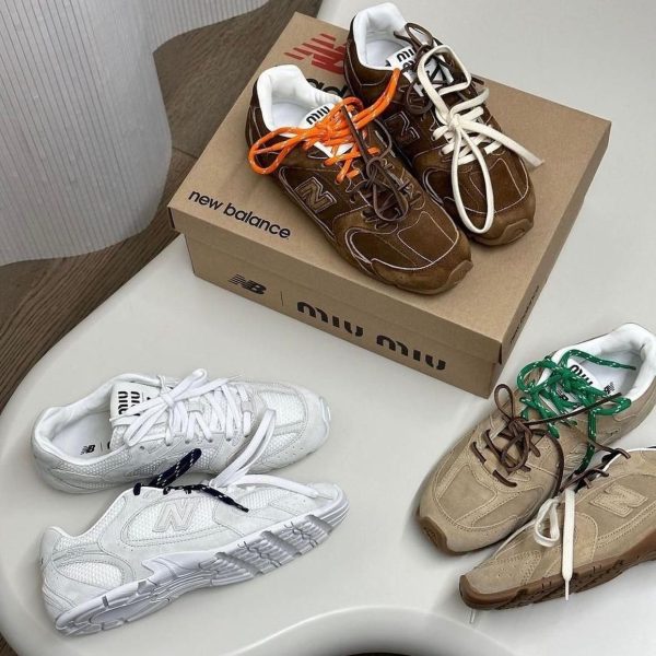 Brown suede New Balance sneakers with orange and white laces resting on a beige New Balance shoebox on a white table.