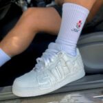 White low-top sneaker with oversized raised letters on the side, worn with white ribbed socks featuring red flowers.