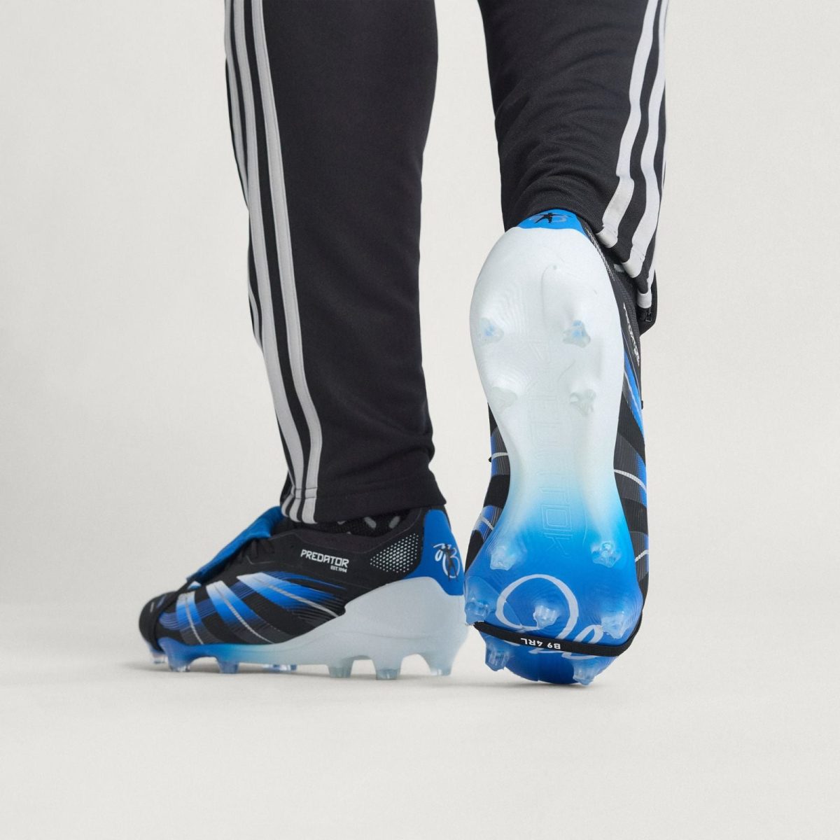 Close-up of a soccer player's legs in black pants with white stripes, wearing blue-and-black soccer cleats with studs showing on the ground.