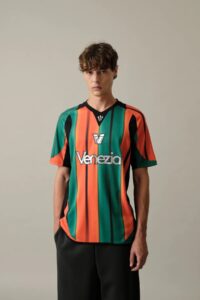 Young man modeling a bold vertical striped Venezia soccer jersey in green, orange, and black against a plain background