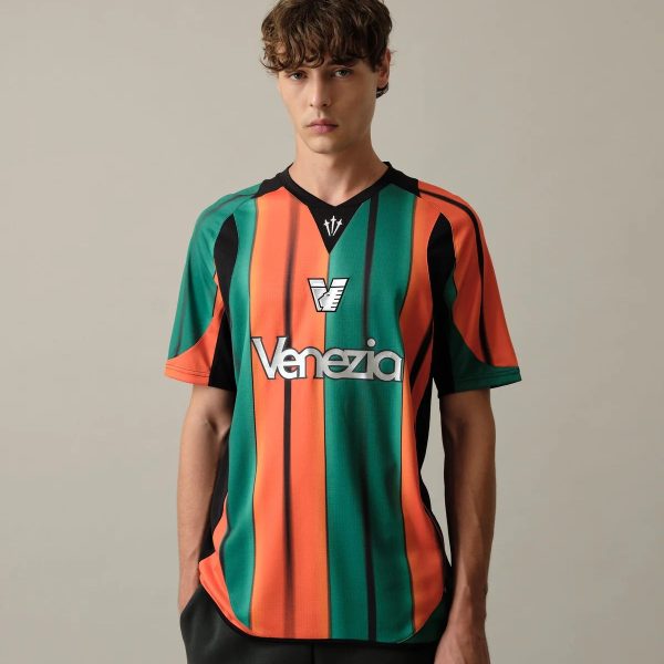 Young man modeling a bold vertical striped Venezia soccer jersey in green, orange, and black against a plain background