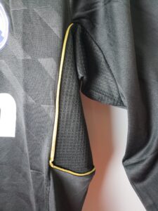 Close-up of a dark textured blazer with a thin gold piping along the front edge and sleeve, showing rolled cuffs.