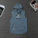 Blue sleeveless hooded top with a front pocket and black drawstrings, brand logo near the hem; sneakers in a wire basket to the right and magazines to the left.