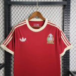 Red Adidas sports jersey on a hanger with white stripes on the shoulders and cream cuffs; crest on the left chest and white Adidas logo on the right.