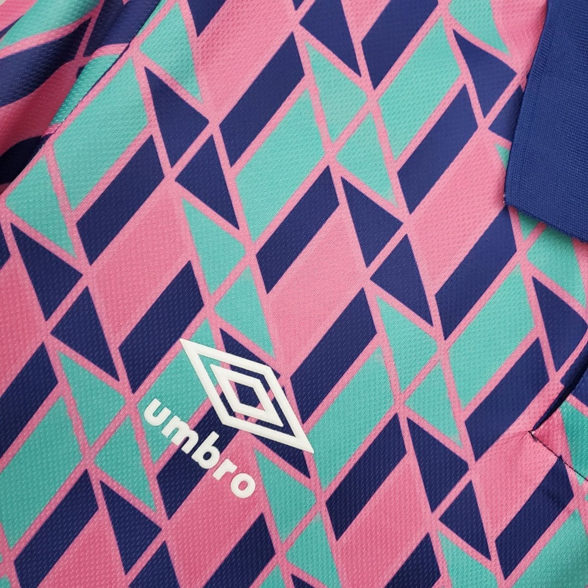 Close-up of a bright geometric fabric pattern with pink background and teal and dark blue diamonds; a partial white logo is visible in the corner.