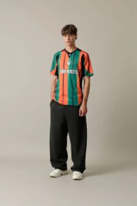 Male model wearing a green, orange, and black striped Venezia jersey with black pants and white sneakers, posing in a neutral studio backdrop.