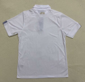 White polo shirt laid flat showing back with 'URUGUAY 1924-2024' and subtle vertical stripe detail; sleeves have dark stripes on ends.