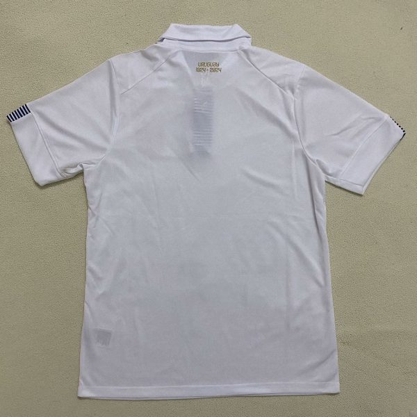 White polo shirt laid flat showing back with 'URUGUAY 1924-2024' and subtle vertical stripe detail; sleeves have dark stripes on ends.