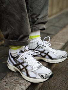 Close-up of white running shoes with brown overlays and mesh panels, worn with white socks and rolled-up gray pants on a stone step in an urban setting.