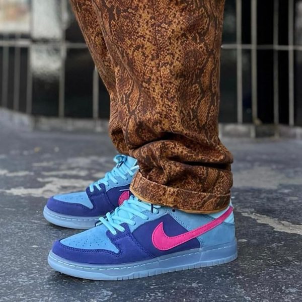 Blue Nike sneakers with a pink swoosh and brown patterned pants standing on a paved street surface.