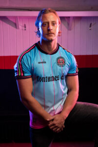 02581803-0201-4F91-A1B8-06787EB54FDA | UAE SPORT STORE Man with blond hair and mustache seated, wearing a light blue football jersey with pink stripes and a crest, hands clasped in front, pink and blue stage lighting behind him.