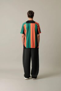 Person standing with back to camera wearing a vertical multicolor striped shirt (green, orange, black) and black pants, white sneakers visible.
