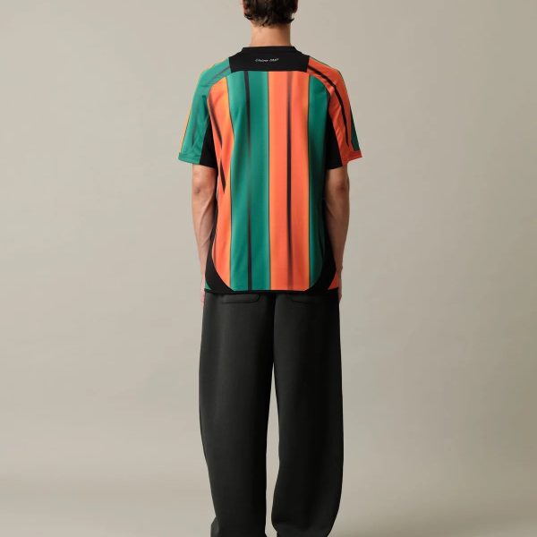 Person standing with back to camera wearing a vertical multicolor striped shirt (green, orange, black) and black pants, white sneakers visible.