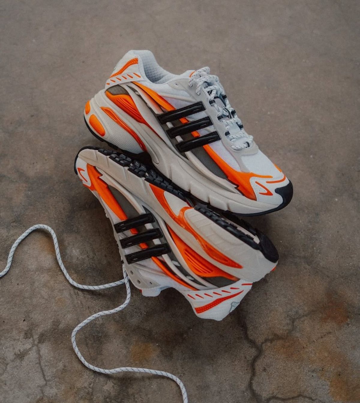 Pair of white athletic sneakers with bright orange accents stacked on a gray concrete surface, shoelaces loose.