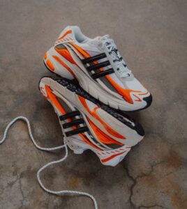 Pair of white athletic sneakers with bright orange accents stacked on a gray concrete surface, shoelaces loose.