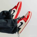 Red and white sneakers with black laces and a black swoosh, paired with cuffed dark jeans against a light background.