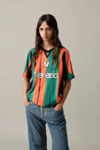 Young woman wearing a colorful vertical-striped Venezia jersey (orange, green, and black) with a Venezia logo, hands in pockets, neutral background.