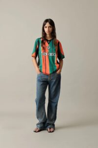 Young woman stands against a neutral wall wearing a bold, multicolored striped Venezia jersey with blue jeans and sandals.