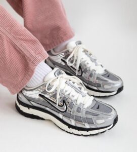Pair of silver and gray Nike sneakers with white laces worn with white socks and pink corduroy pants, close-up of footwear on a light background.