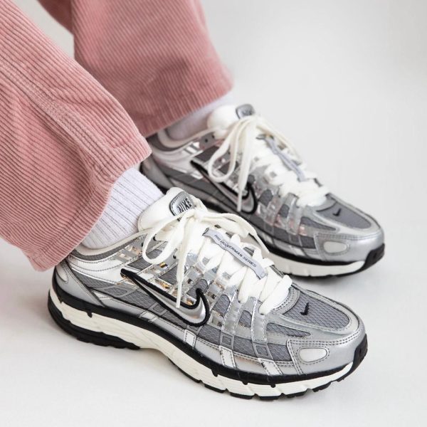 Pair of silver and gray Nike sneakers with white laces worn with white socks and pink corduroy pants, close-up of footwear on a light background.