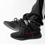 Black Yeezy Boost 350 sneakers with red SPLY-350 lettering, being worn with jogger pants (side view).