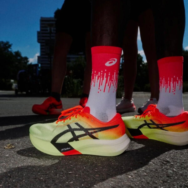 Close-up of neon yellow–orange running shoes with white-to-red gradient socks on asphalt, runners in the background ready to run.