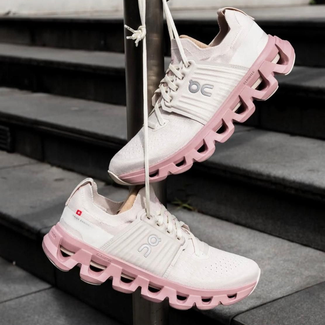 05ae4980-47a5-4d16-92e3-af7a9f0e6c50 | UAE SPORT STORE White sneakers with pink soles hanging from a metal pole on concrete stairs beside a railing.