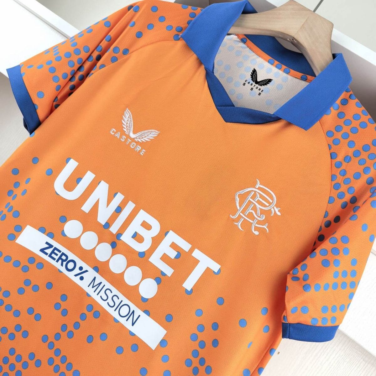 Close-up of an orange athletic jersey with blue polka dots, blue collar and cuffs, featuring Castore branding and a team crest.
