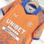 Close-up of an orange athletic jersey with blue polka dots, blue collar and cuffs, featuring Castore branding and a team crest.