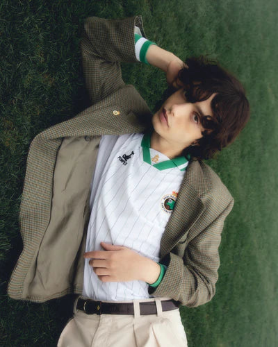 06-2web_400x_c6006f86-5af6-4ae9-a235-f9e3d59fa7db | UAE SPORT STORE Person lying on a grassy field, wearing a white pinstripe athletic jersey with green trim and a tan blazer, hand resting on their head.