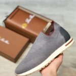 Grey knit slip-on sneaker with white sole and black heel cap, held near brown shoeboxes in the background