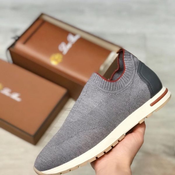 Grey knit slip-on sneaker with white sole and black heel cap, held near brown shoeboxes in the background