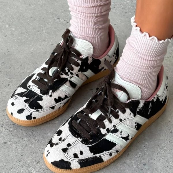 Child wearing pink ribbed socks and white-and-black cow-print sneakers with brown laces on a concrete surface.