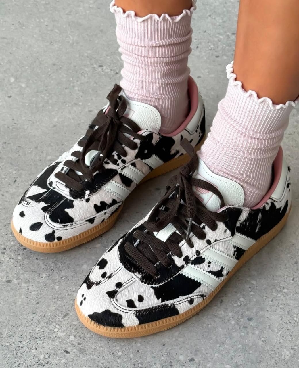 0659aabe-11ab-4b2f-a9b5-b5c8f18f9474 | UAE SPORT STORE Child wearing pink ribbed socks and white-and-black cow-print sneakers with brown laces on a concrete surface.
