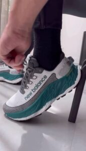 Close-up of a hand tying the lace on a white and teal New Balance running shoe, black sock visible, chair leg in the background.