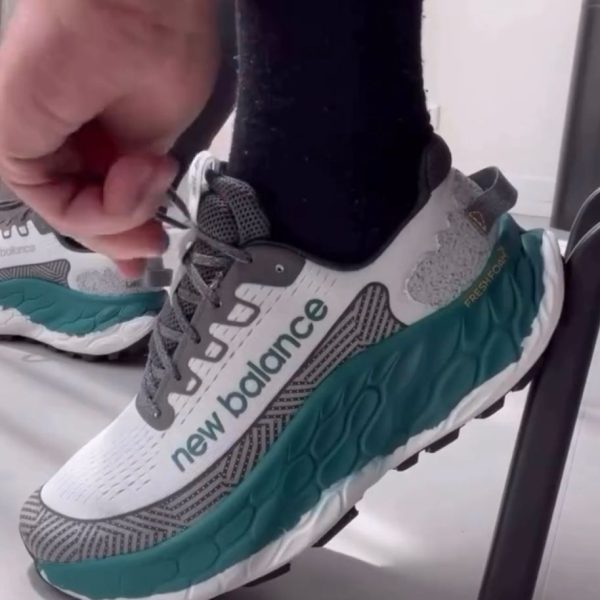 Close-up of a hand tying the lace on a white and teal New Balance running shoe, black sock visible, chair leg in the background.