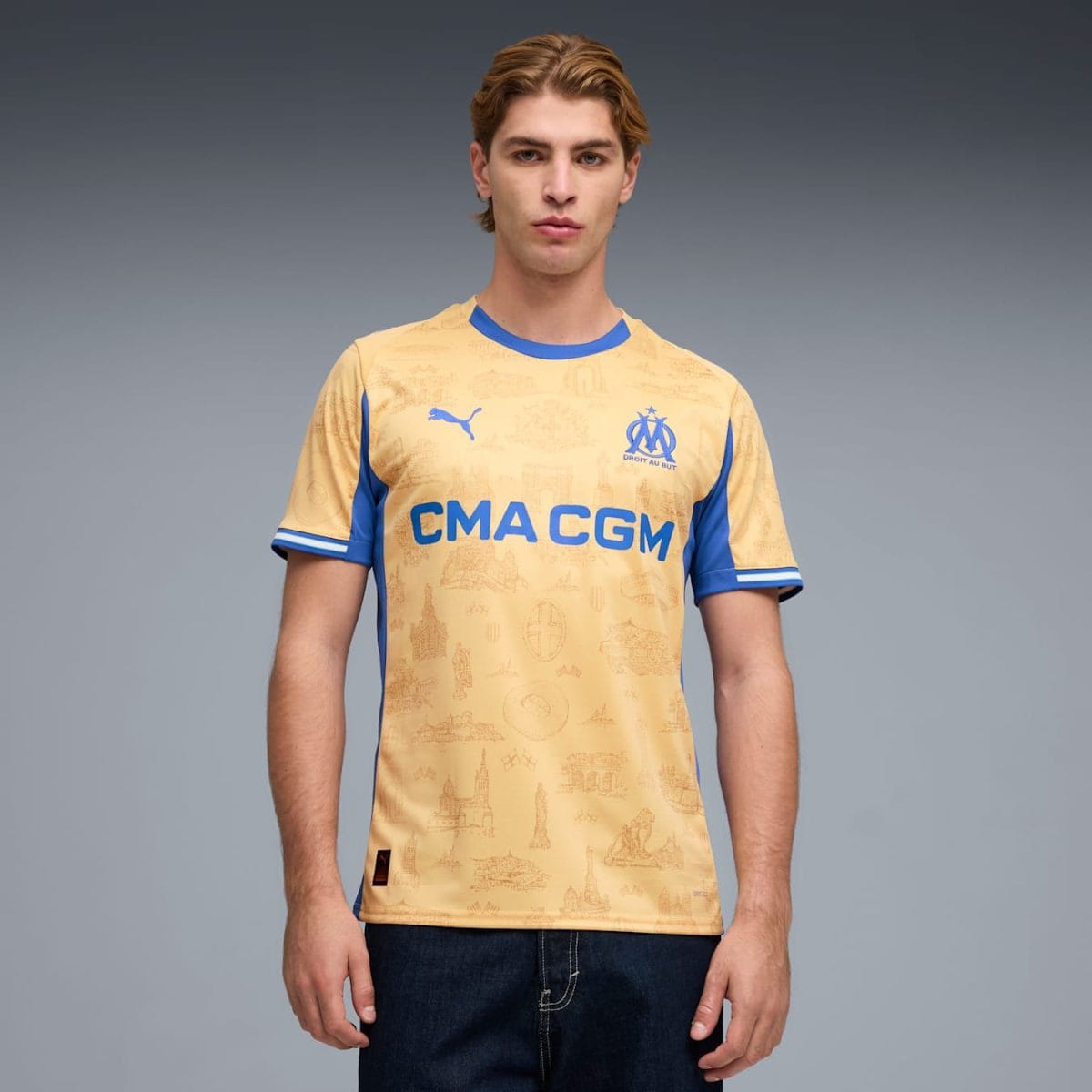 Young man wearing a beige Olympique de Marseille football jersey with blue trim and CMA CGM sponsor, standing against a gray gradient background