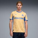 Young man wearing a beige Olympique de Marseille football jersey with blue trim and CMA CGM sponsor, standing against a gray gradient background